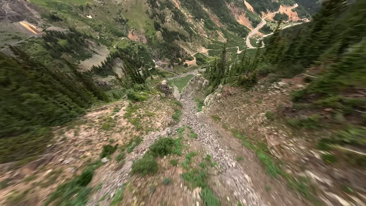 A shot from an FPV drone racing through a steep mountain couloir, between cliffs, overlooking the Million Dollar Highway (Red Mountain Pass) in the San Juan Mountain Range of Colorado in the Summer