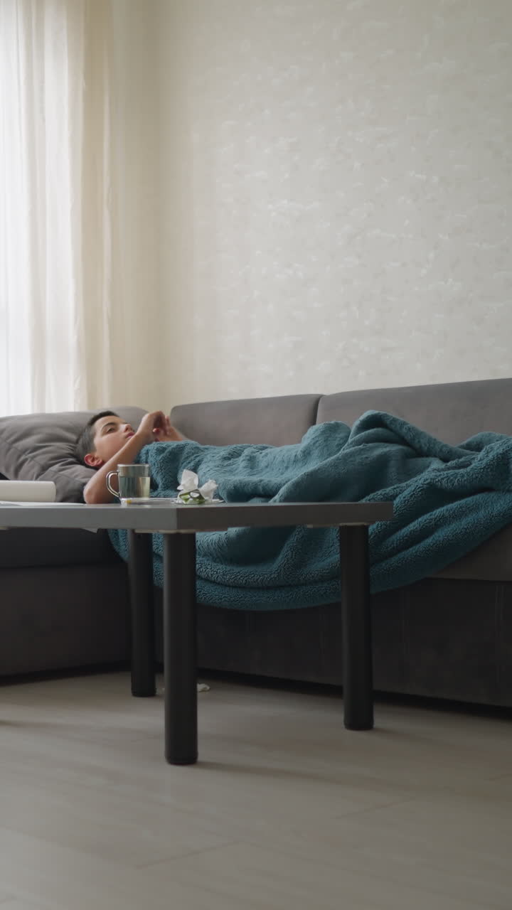 Child lying on couch covered with blanket adjusting for comfort with glass cup on table in living room, peaceful indoor setting with soft natural light coming through window