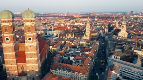 Paso-Aéreo-Cerca-De-Las-Torres-De-La-Maravilla-Arquitectónica,-Munich-Frauenkirche,-Rathaus-La-Perspectiva-Aérea-Revela-El-Paisaje-Urbano-De-Munich-En-El-Horizonte-Durante-El-Sol-De-ángulo-Bajo