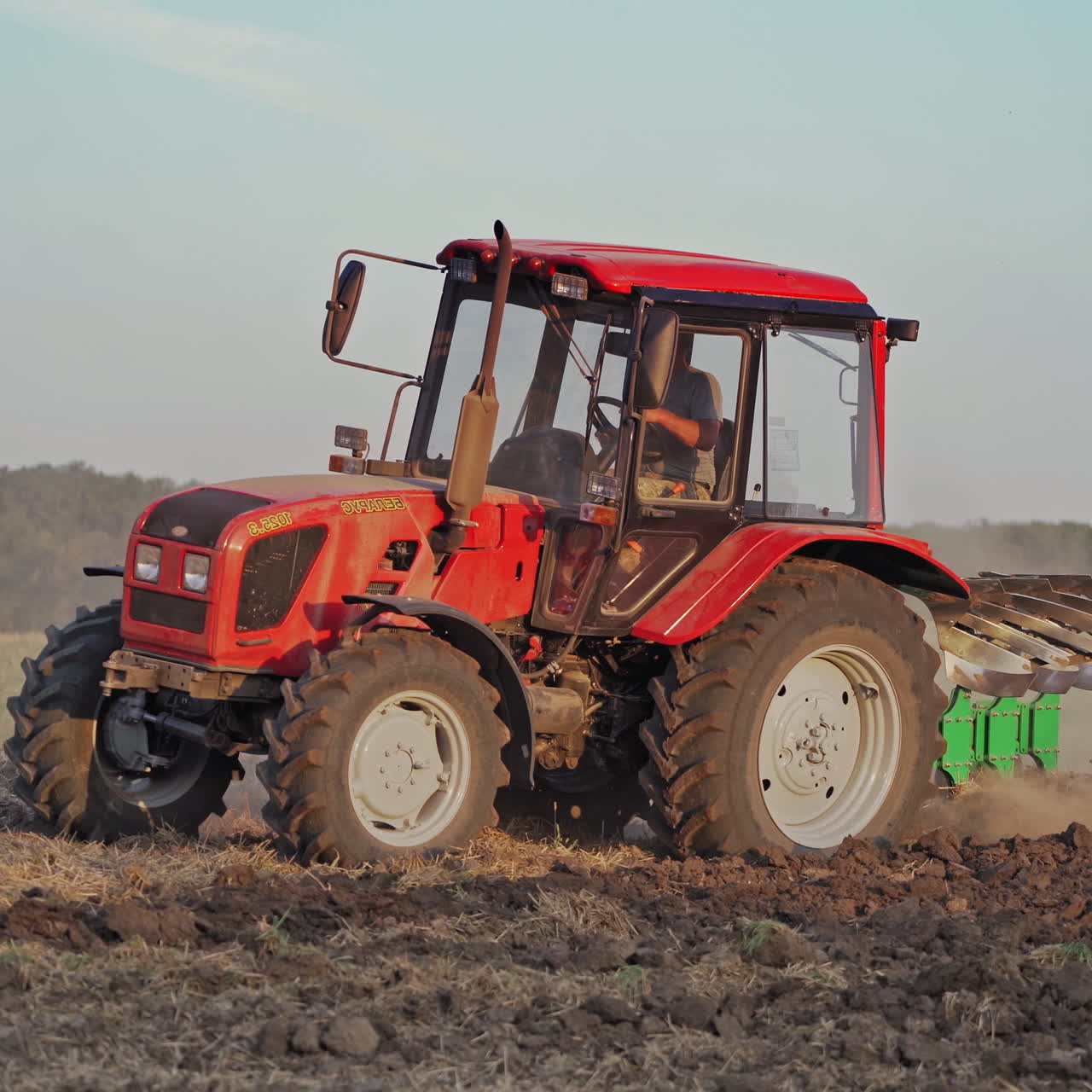 Side view of a red tractor cultivating the soil on the field. Farmer in tractor prepares land for sowing in farmlands outdoors.