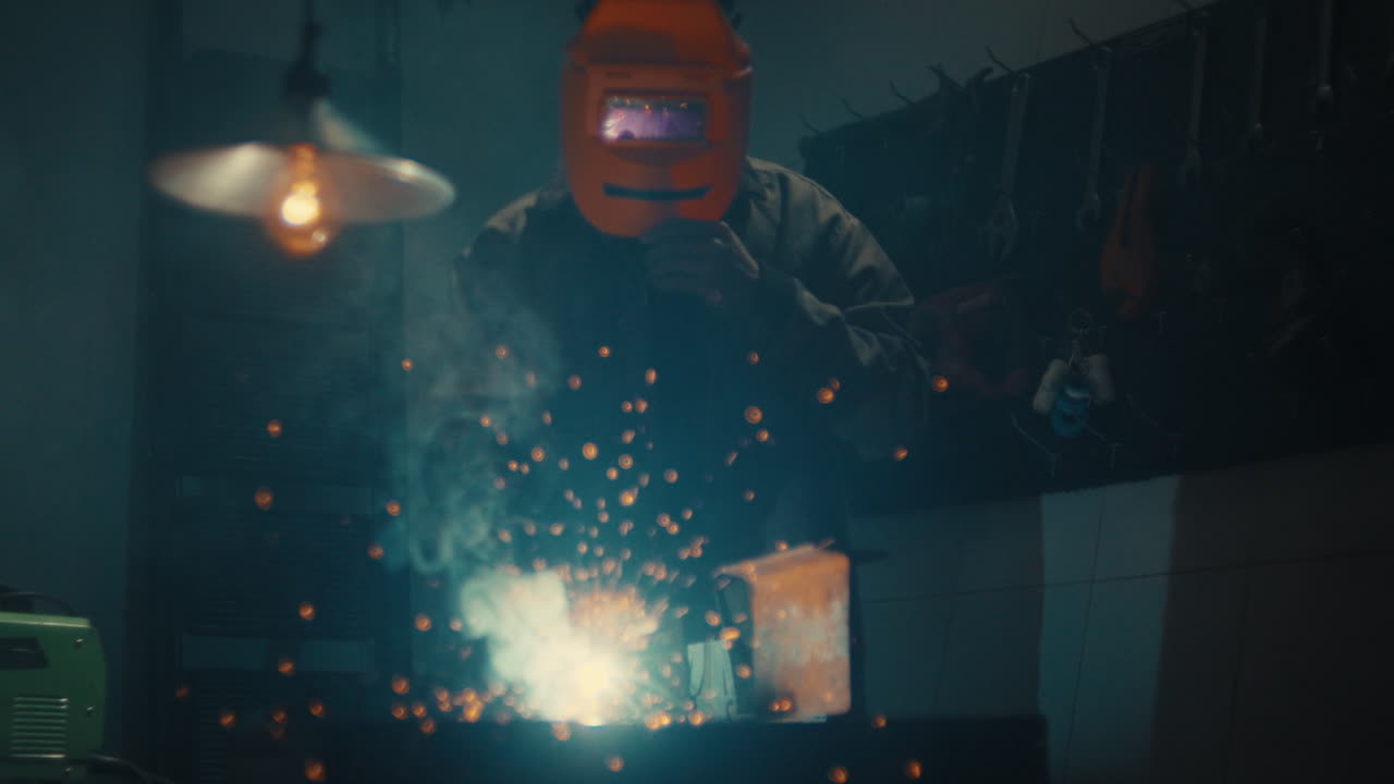 Welding metal with bright sparks flying in dark workshop, slow motion glow with man wearing welding safety suit