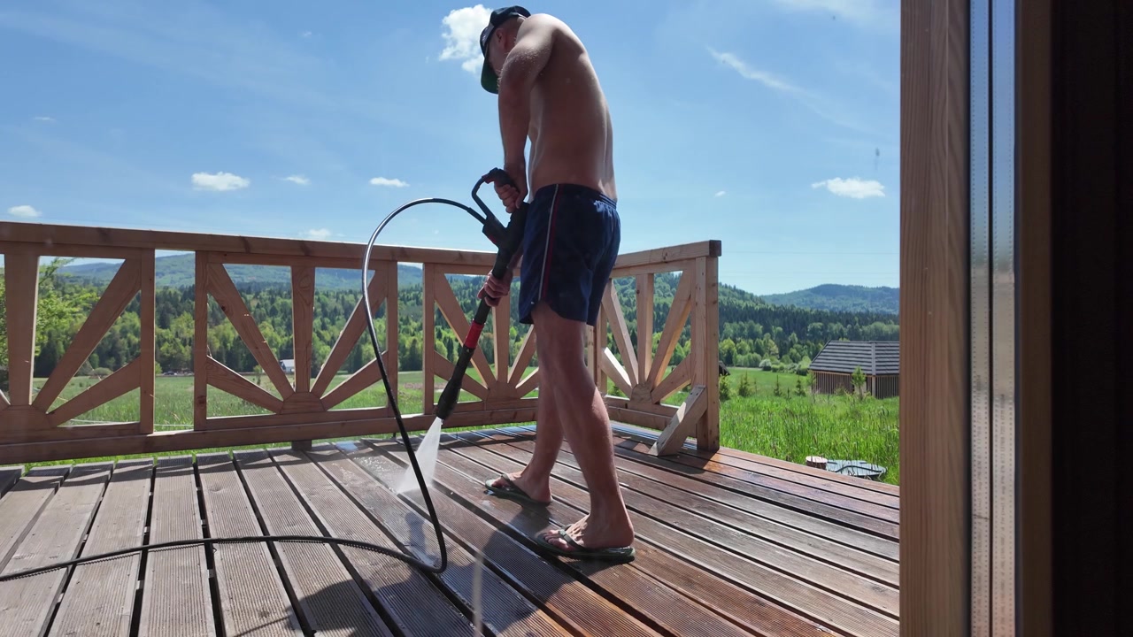 Man cleaning a wooden deck with a pressure washer outside a rustic log house in a mountain landscape on a sunny summer day.