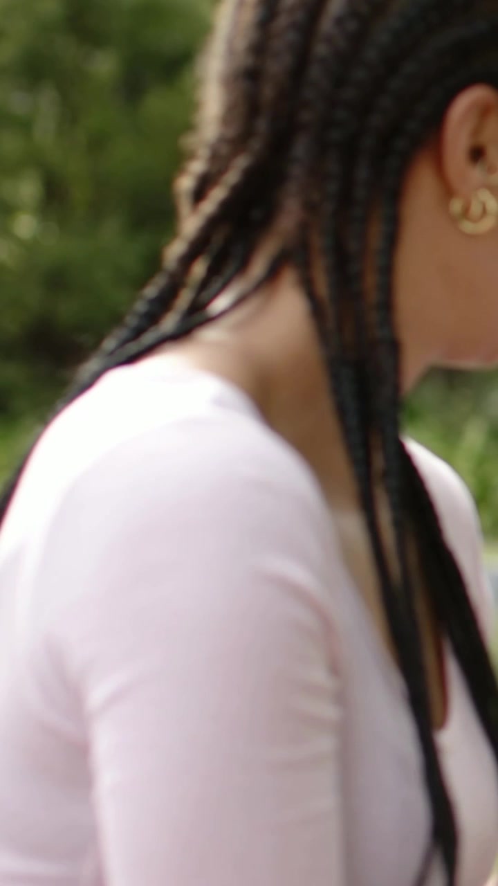 Vidéo verticale : Entendant un appel, femme se retournant et souriant dans un parc, avec des tresses, un haut rose et des créoles dorées.