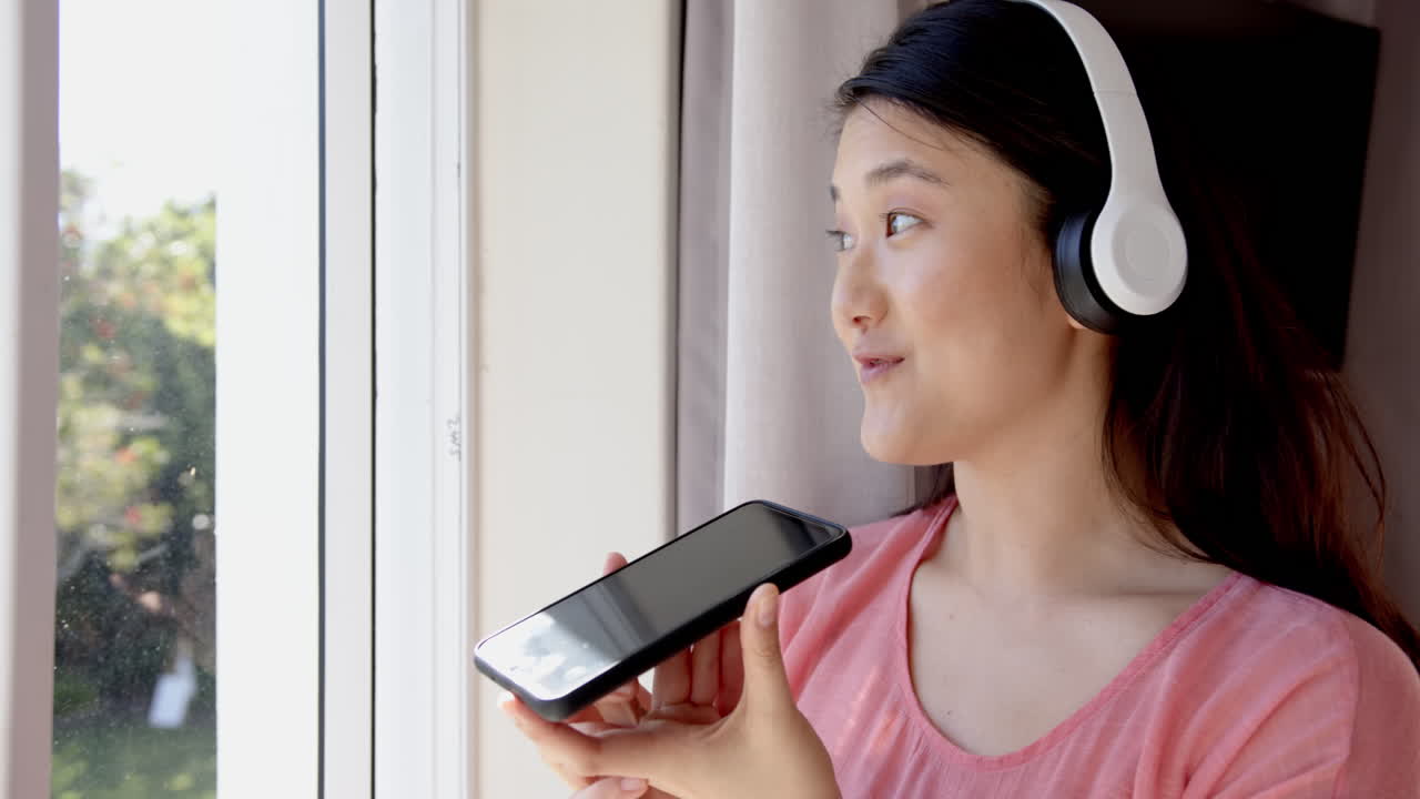 Using smartphone and wearing headphones, woman talking and looking out window