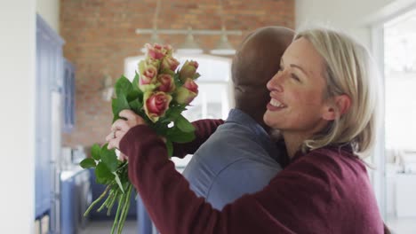 Feliz-Pareja-Diversa-De-Alto-Nivel-En-La-Sala-De-Estar-Abrazándose,-Sosteniendo-Flores