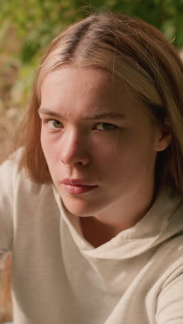 young adult seated on ground under tree, hand resting on knee, looking up contemplatively, dry foliage and earthy tones in background
