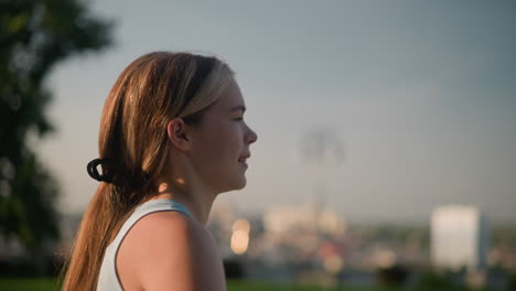 side view of young lady smiling as sunlight reflects on her face while she moves gracefully like one skating, hair tied back, blurred background with natural glow, trees, and an unclear object