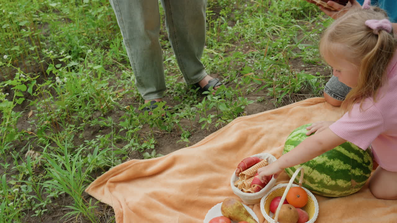 흰색 옷을 입은 남매가 자작나무 아래 오렌지색 담요 위에서 피크닉 바구니를 정리하고 있어요. 오빠는 빵을 놓고 여동생은 사과를 잡으려고 손을 뻗고 있으며 부모는 근처에 서 있습니다. 수박, 고리버들 쟁반, 소박한 분위기