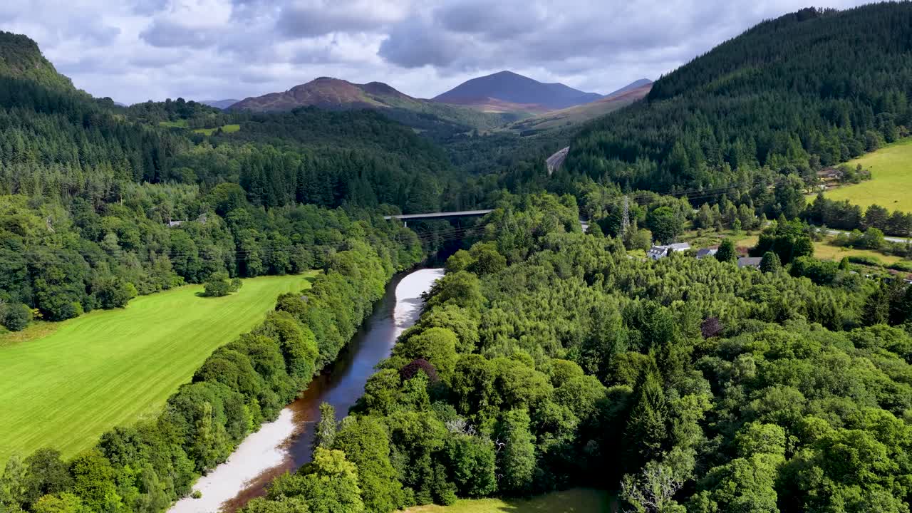 Drone glides above lush green valley, river, and forested hills under partly cloudy daylight