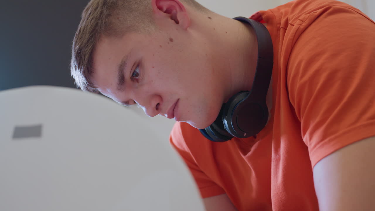 Young technician wearing orange shirt and black headphones leans forward with intense focus and concentration working attentively on unseen task showing dedication
