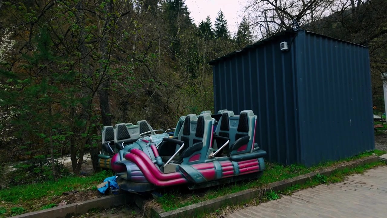 Pembe ve mavi oturma düzeneklerine sahip, koyu mavi metal bir kulübenin yanına yerleştirilmiş, ilkbahar yeşillikleri ve ormanla çevrili terk edilmiş bir lunapark oyuncağı.
