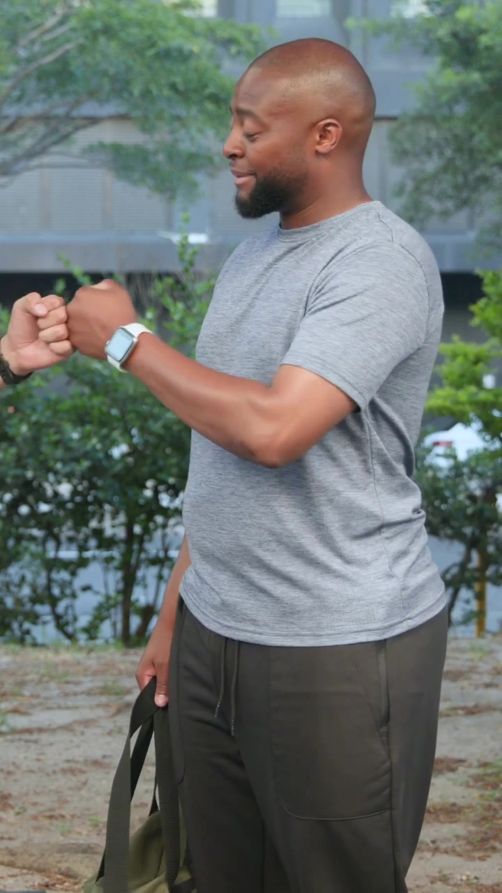 Vertical video: Checking watch, man greeting arriving friend in park with bump, man with duffel