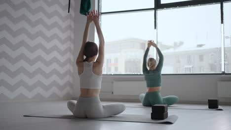 Two-women-do-back-exercises-together-in-front-of-a-window-in-the-fitness-room.-Two-women-perform-a-stretch-and-warm-up-of-the-back-together-with-the-instructor.