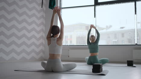 two women do back exercises together in front of a window in the fitness room. two women perform a stretch and warm-up of the back together with the instructor.