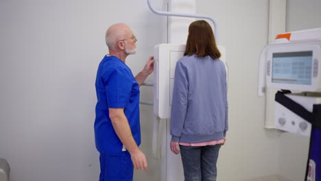 A-young-brunette-girl-at-an-appointment-with-a-doctor-in-a-blue-uniform-with-a-gray-beard-during-fluorography-in-a-modern-office-in-a-clinic.-Brunette-girl-undergoing-fluorography-in-a-modern-clinic-under-the-supervision-of-a-doctor