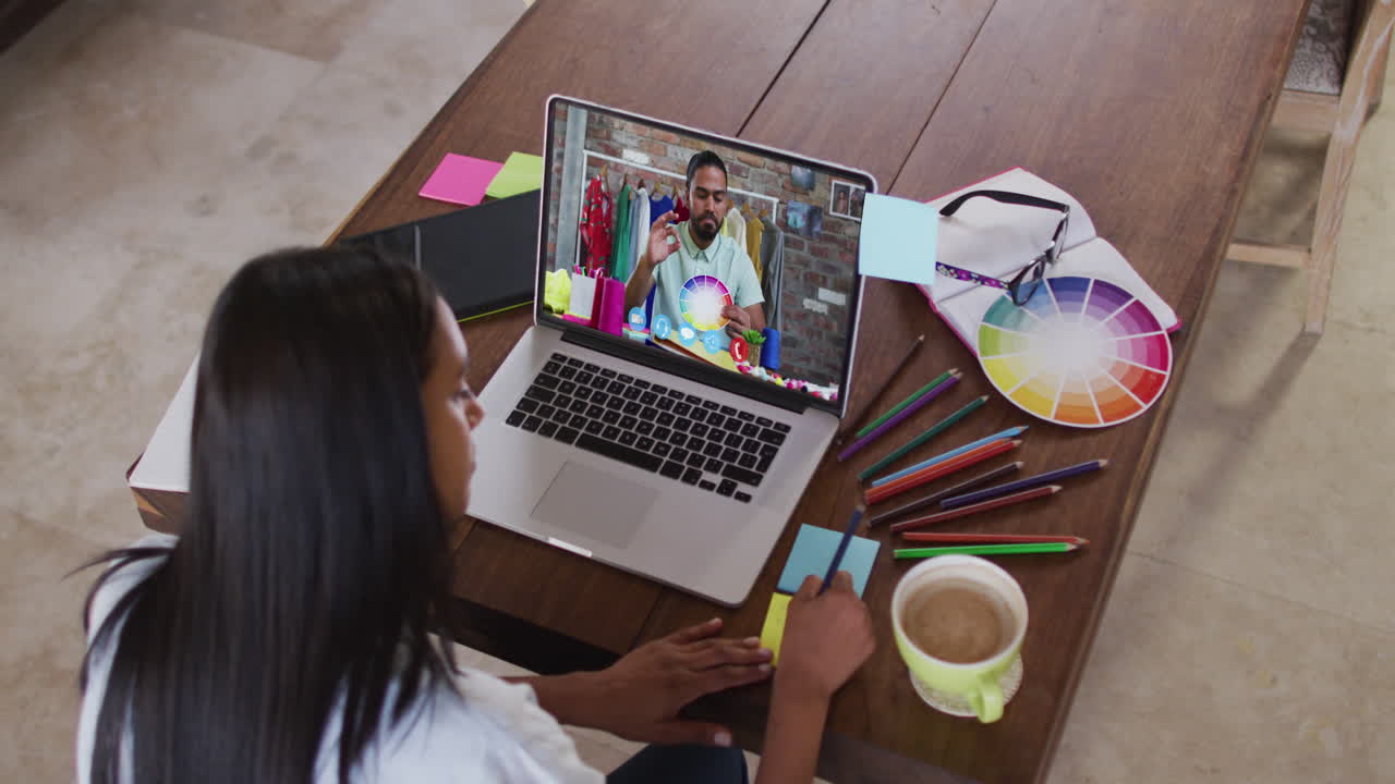mujer afroamericana tomando notas mientras tiene una videollamada con un colega masculino en una computadora portátil en casa
