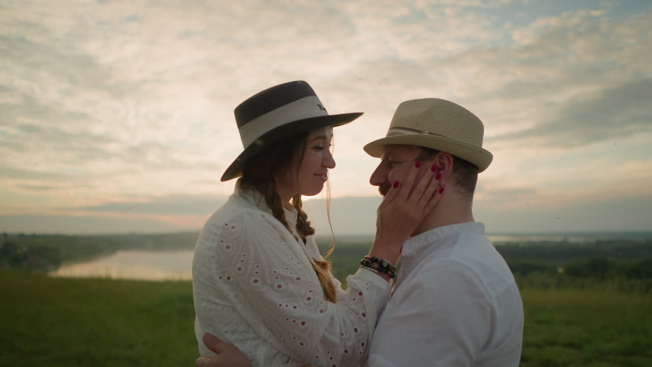색 드레스를 입고, 모자와 팔를 착용 한 한 여성은 해가 지는 동안 호수 의 잔디에서 남편의 얼굴을 사랑스럽게 만질 때 따뜻하게 미소 짓고, 남편은 색 셔츠와 모자를 입고 있습니다.