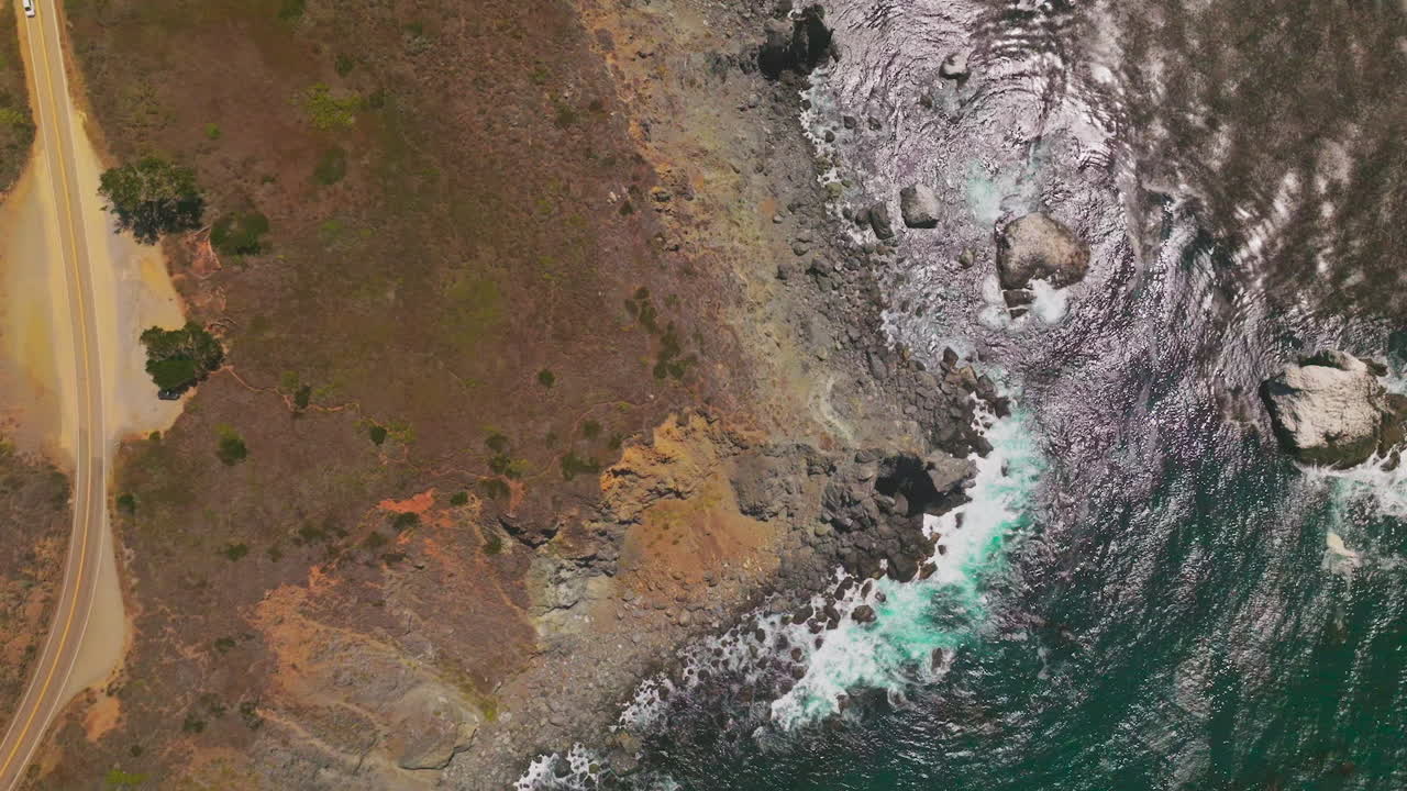 Sparkling in the sun water of the ocean washing the rocks at California shore. Drone footage descending over the rocky coastline with highway on it.