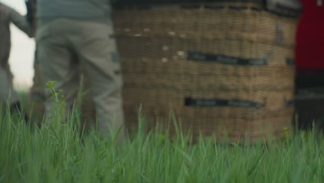 close up of people dragging wicker basket across tall grass toward paramotor trailer at dusk under soft cloudy sky, focus on textured weave and teamwork effort