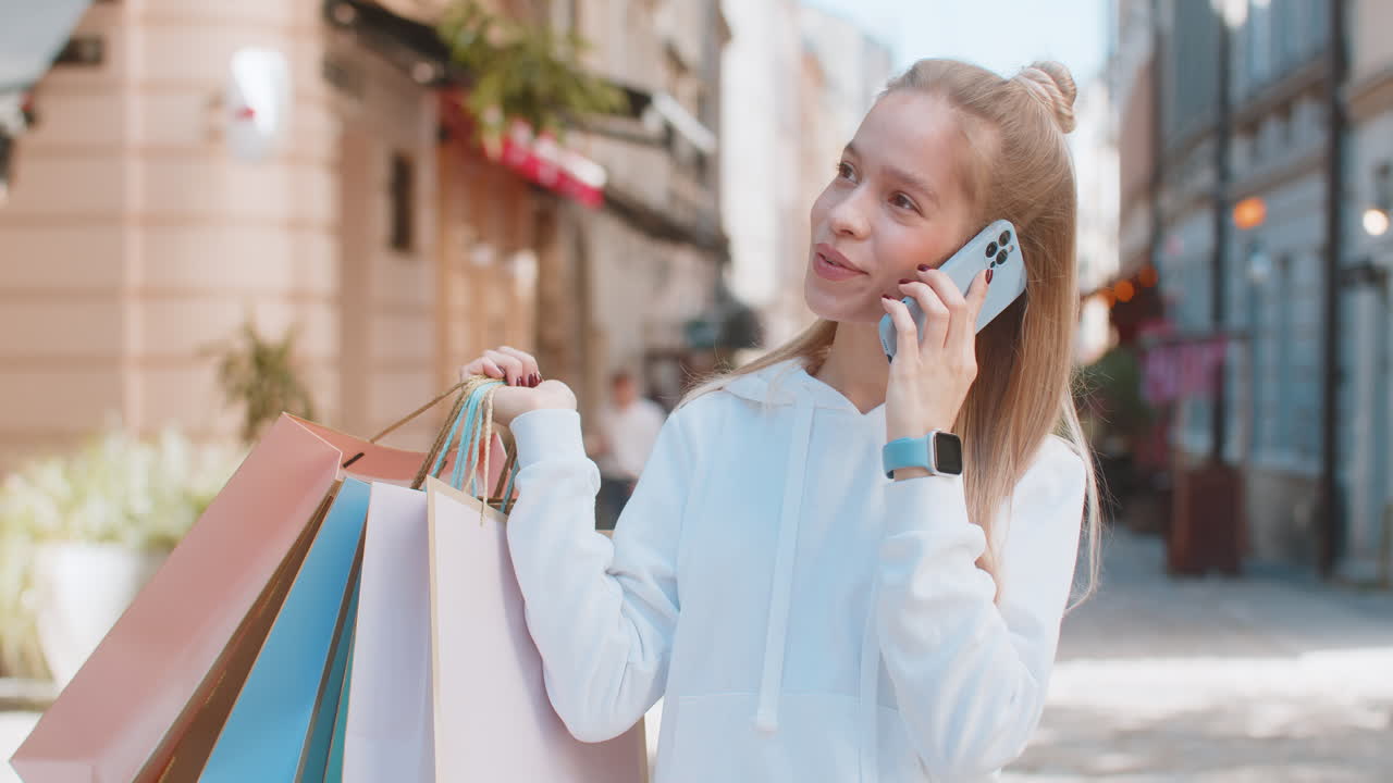 joven feliz adolescente hablando en un teléfono inteligente de pie en la calle de la ciudad sosteniendo bolsas de compras