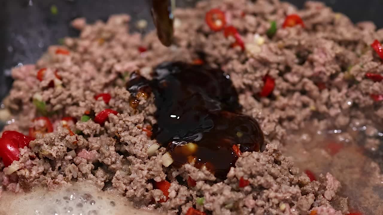Ein Koch gibt eine dicke, dunkle Soße zu brutzelndem Hackfleisch und roten Paprika in einer Pfanne hinzu