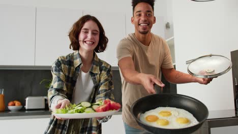 Portrait-of-a-happy-young-man-with-Black-skin-in-a-cream-T-shirt-and-a-young-adult-girl-with-a-bob-hairstyle-in-a-plaid-shirt-who-holds-in-her-hands-a-salad-and-a-fried-egg-while-preparing-breakfast-in-a-modern-kitchen