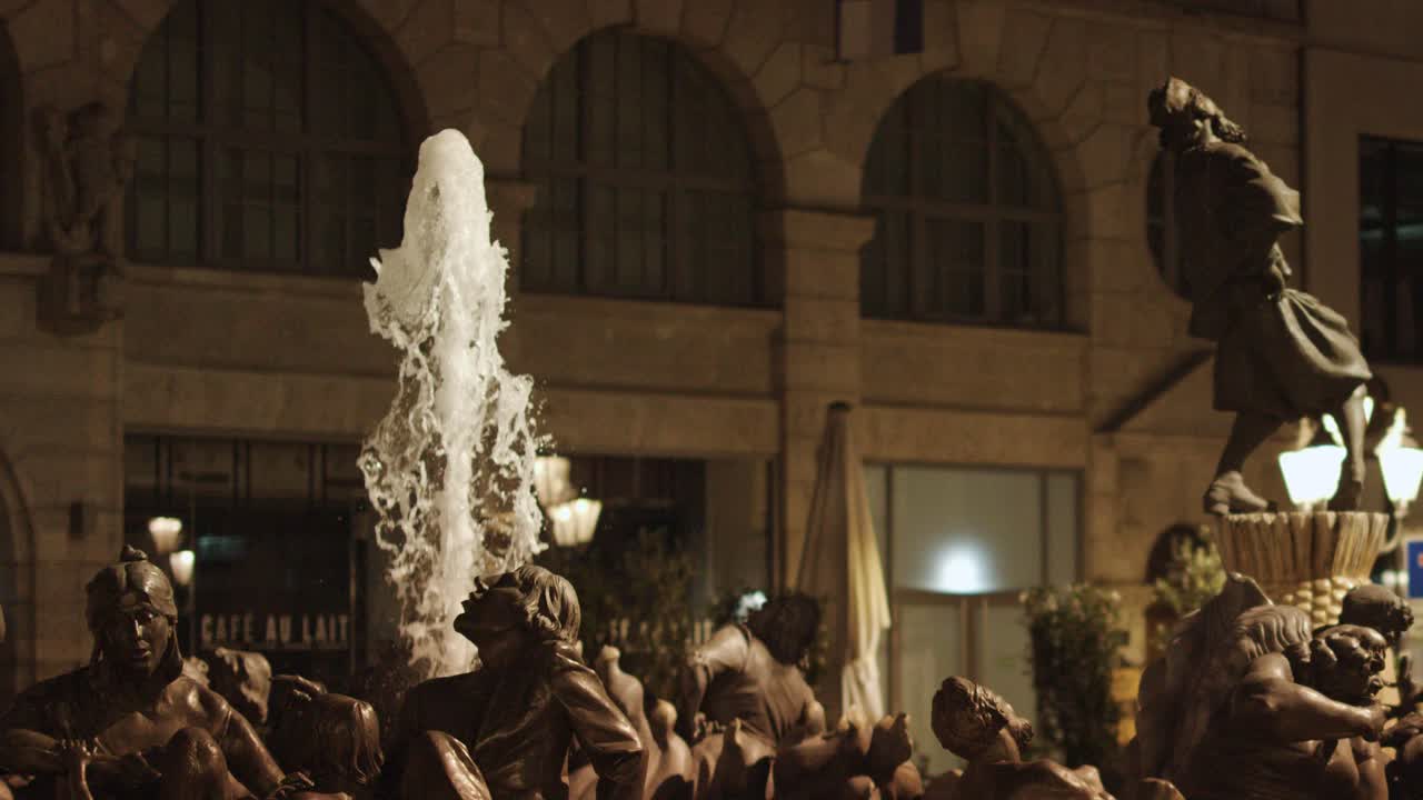 Fontana con statua in bronzo che erutta acqua di notte, illuminata da caldi lampioni, con sfondo urbano