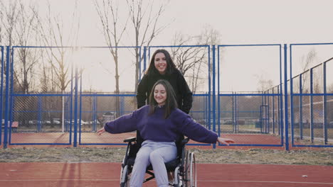 young woman running while pushing her disabled friend in wheelchair and having fun together in basketball court