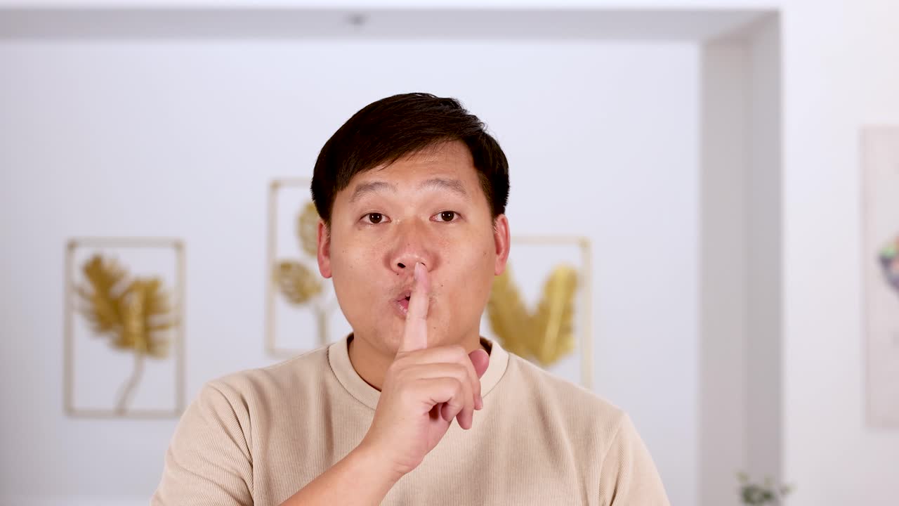 Asian man holds finger to lips in a shushing gesture, facing camera in a well-lit, modern living room with neutral decor and soft lighting
