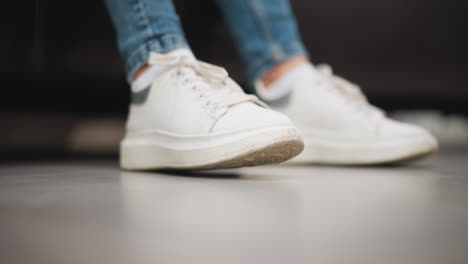 close-up of lady's white canvas sneakers gently tapping her right foot on the floor while seated on a couch wearing blue jeans in a comfortable indoor setting