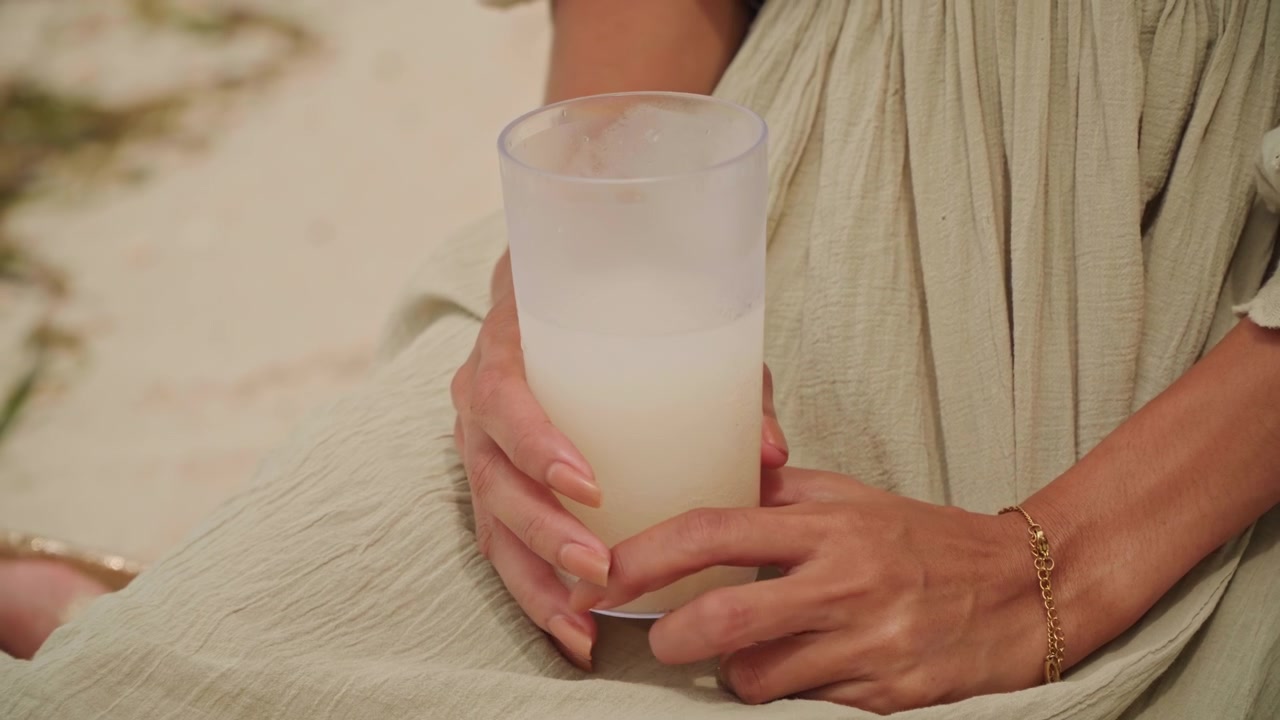 Donna che tiene un bicchiere satinato sulla spiaggia, primo piano delle mani con braccialetto d'oro e vestito di lino, bevanda lattiginosa chiara, sfondo di sabbia soffice, calda luce del tramonto, atmosfera estiva rilassata, momento di vita spontaneo