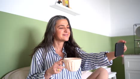 happy mature with coffee having video call in bedroom