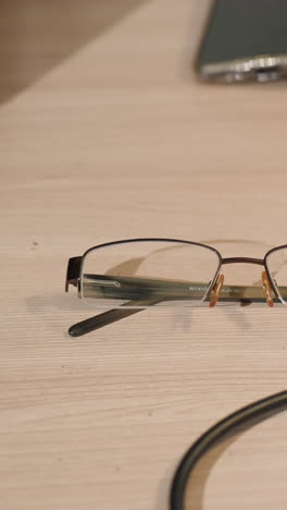 una vista de cerca de alguien con la mano derecha usando un guante negro alcanzando un par de gafas en un escritorio de madera, con teléfono en la mesa y otros equipos