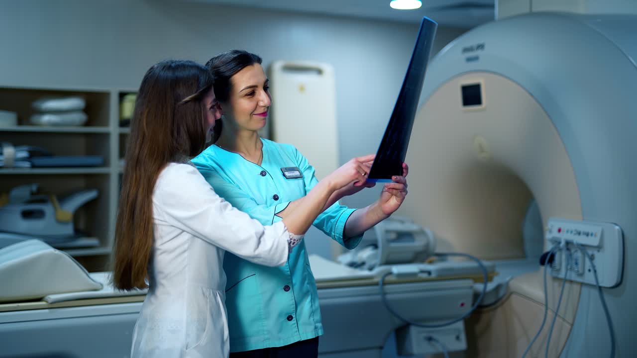 Medical workers discuss the x-ray photograph. Two female specialists in medical uniform examining x-ray photo on the background of new equipment in clinic.