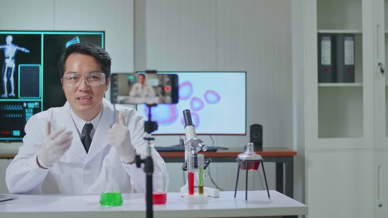 Asian Scientist Man Recording Webinar, Using Smartphone On Tripod, Sitting At Desk In Office