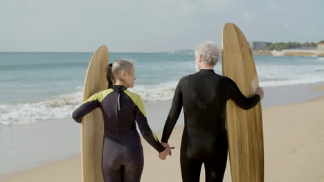 vista trasera de una pareja mayor en traje de neopreno con tabla de surf de pie en la playa de arena y tomados de la mano