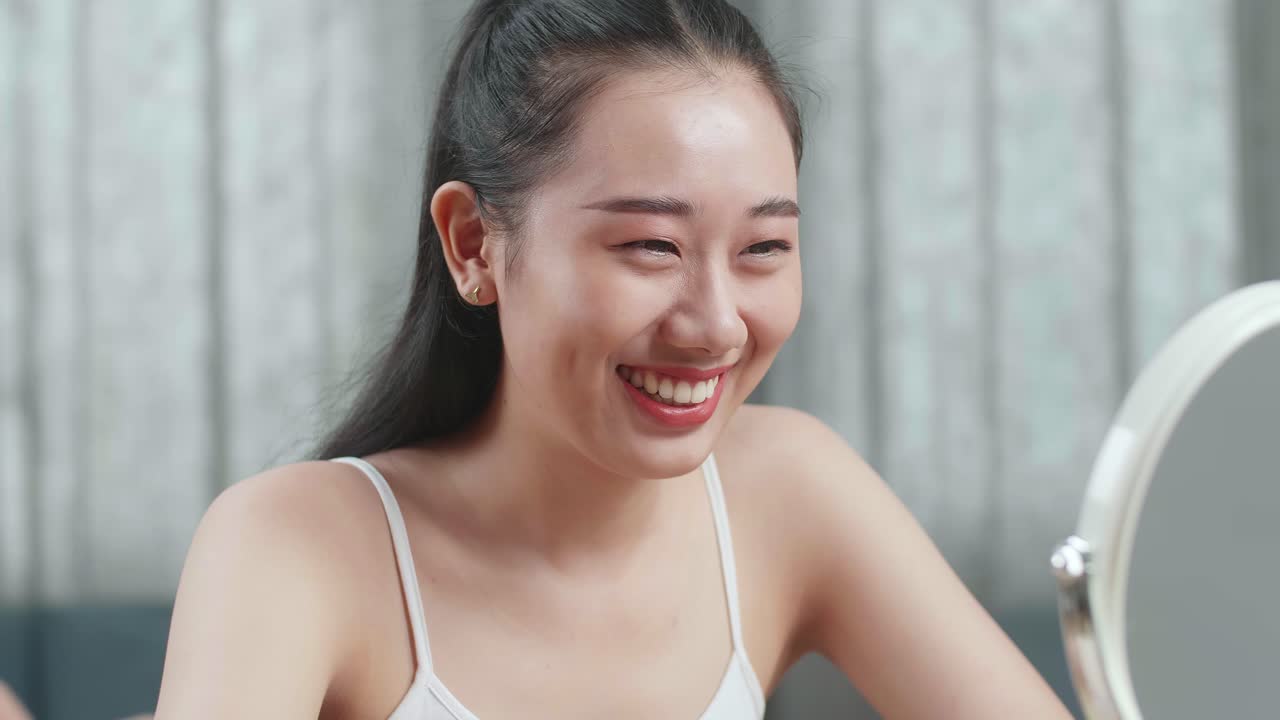 아름다운 젊은 아시아 여성이 거울을 바라보고 집에서 메이크업을 한 후 미소 짓고 있습니다.