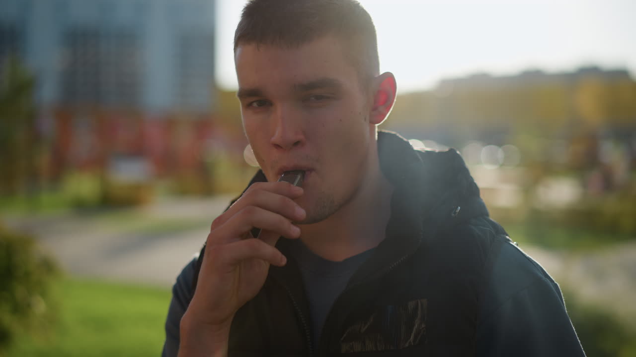 young man in black vest inhales from shisha device and exhales thick white smoke while standing outside in green urban park with modern blurred buildings and evening light in background