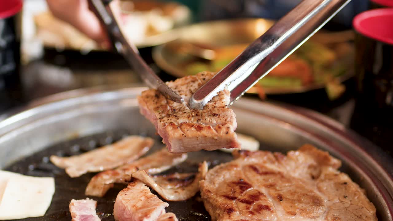 Hand uses tongs and scissors to cut pork belly on hot grill, indoor restaurant lighting