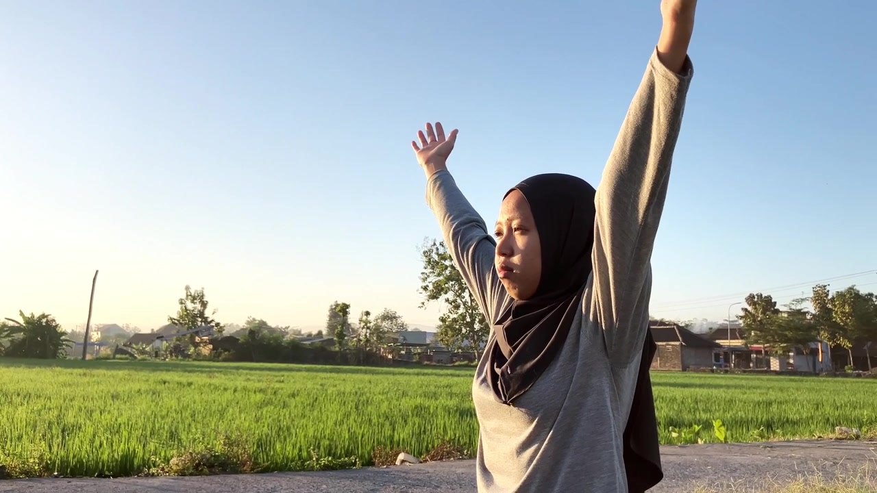 Klip gerak lambat seorang wanita Muslimah berhijab mengangkat tangannya dalam peregangan yoga di luar ruangan. Cahaya matahari pagi yang indah dengan latar belakang sawah hijau subur. Sempurna untuk konsep kesehatan dan kebugaran.
