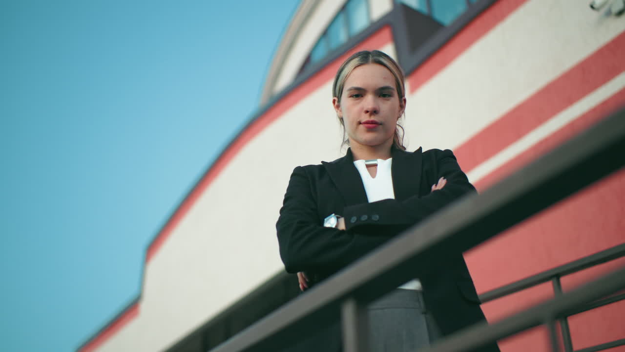Real estate manager in black blazer folds hands while standing confidently in front of organization building with red and white stripes, security camera visible above