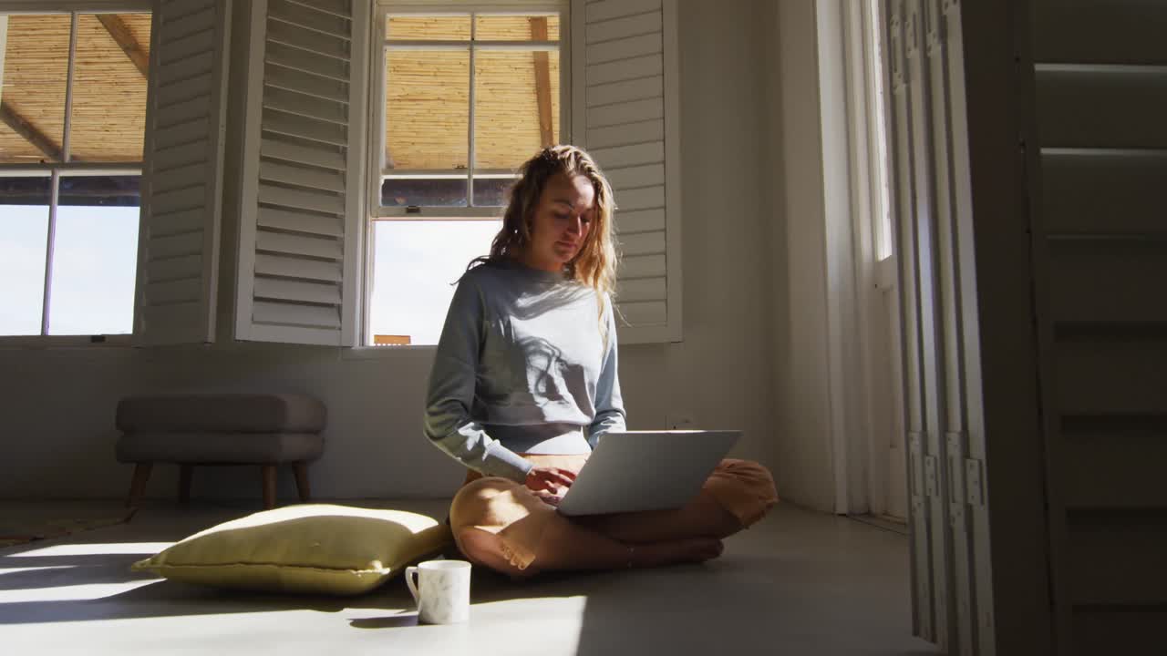 Caucasian woman sitting on floor with cup of coffee using laptop in sunny cottage living room
