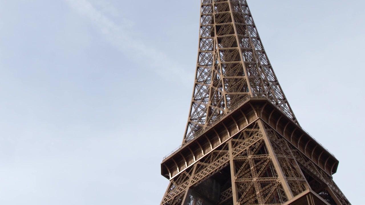 Una telecamera con angolazione bassa si muove verso l'alto lungo la struttura a traliccio in ferro della Torre Eiffel.