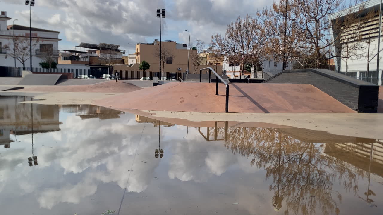 Skatepark vazio cheio de água da chuva, refletindo o céu nublado, as árvores ao redor e os edifícios urbanos, criando uma cena única.