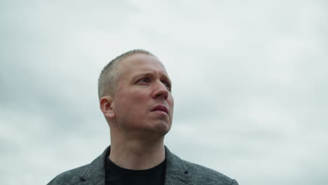 a close view of an aged white man wearing a grey suit and a black polo shirt, turning his attention to the left with a focused expression, with an overcast sky