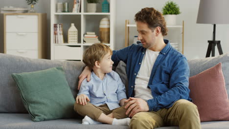 Joven-Y-Apuesto-Padre-Sentado-En-El-Sofá-De-La-Sala-De-Estar-Y-Hablando-Con-Su-Pequeño-Y-Lindo-Hijo-Que-Se-Sienta-A-Su-Lado