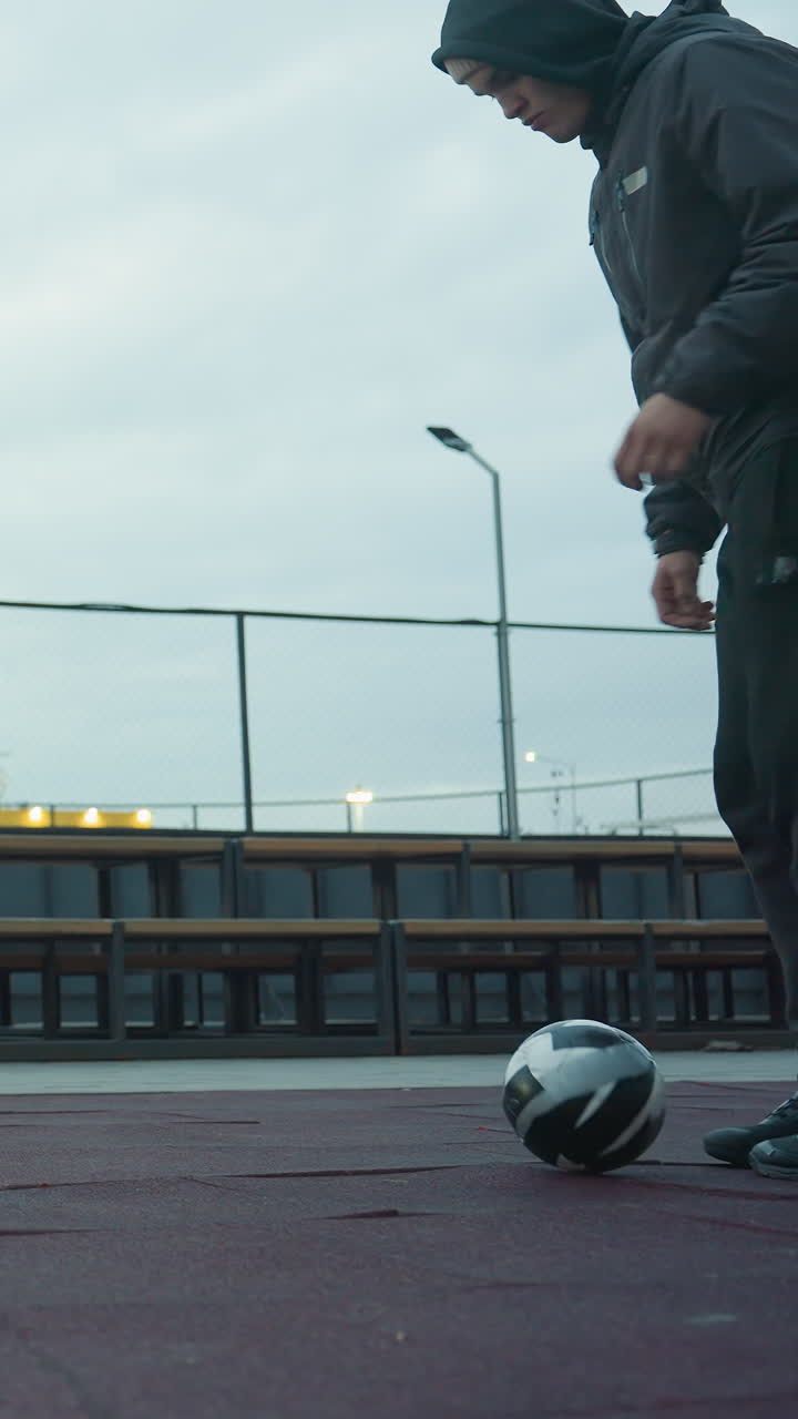 빈 스포츠 경기장의 넓은 으로 벤치, 체인 링크 울타리, 거리 조명과 멀리 떨어진 건물을 포함한 도시 주변에 초점을 맞추고 축구 훈련을 하는 남자