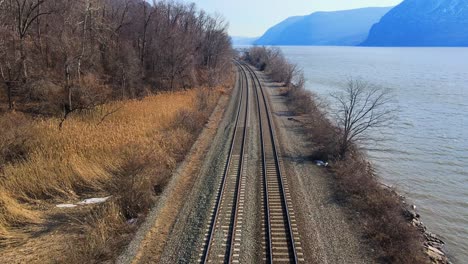 aerial drone footage down railroad train tracks in a river valley with mountains and water in early spring