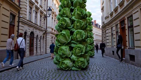 giant lettuce sculpture occupies a narrow street in prague, with people walking by, creating a surreal and thought provoking scene that blends nature and urban environment