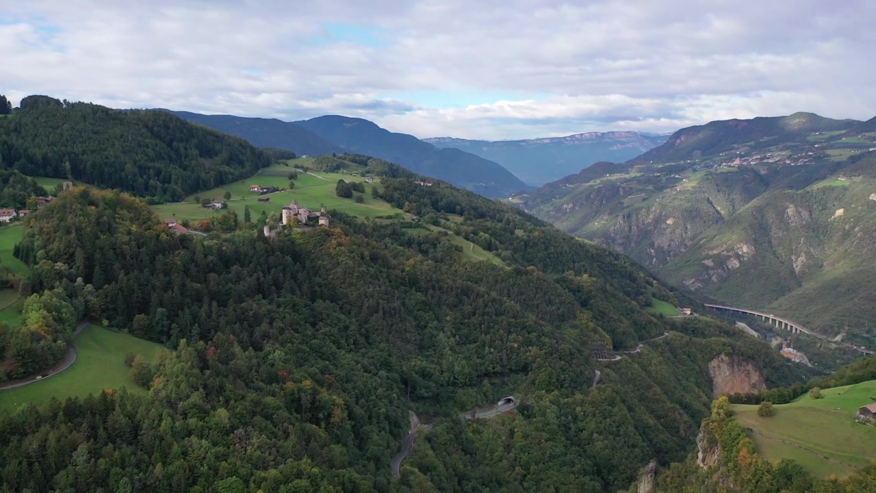 Stunning high-angle aerial drone shot of the historic stone Castel Presule in Italy. Features the medieval fortress perched on a green hill surrounded by lush alpine forests and deep valleys.